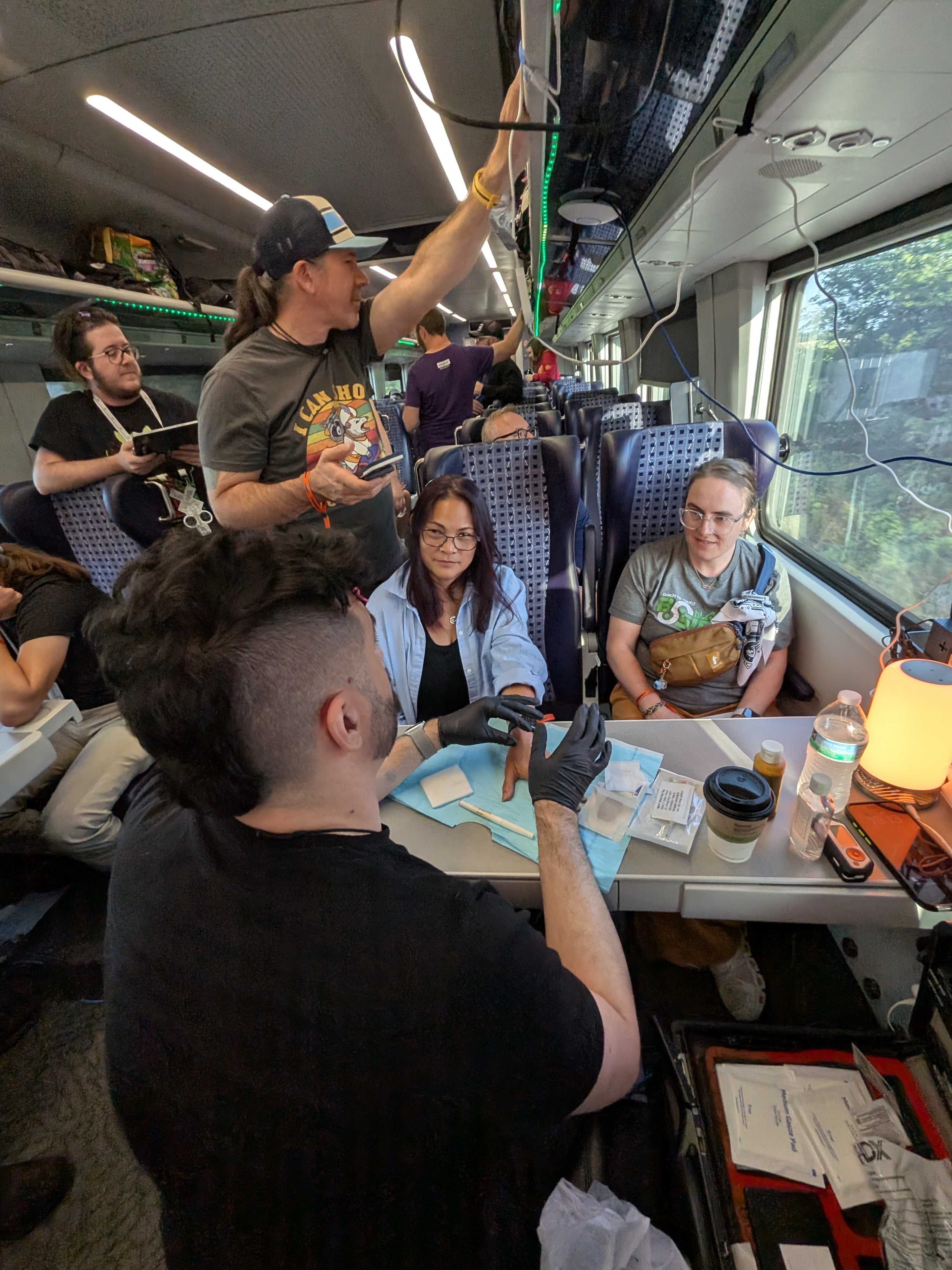 Johnny Xmas installing a biohacking implant for Heidi Potter, supported by Bruce Potter and Jessie Jamieson at a four-seat table on the moving train